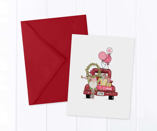 Valentine Gnome holding a wreath over his head in front of a vintage red pickup truck filled with presents