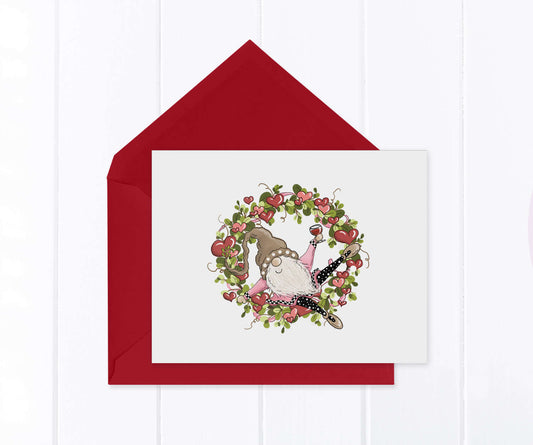 A festive Valentine gnome holding a glass of wine sitting inside a heart and greenery wreath